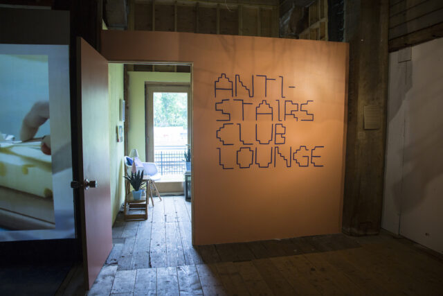  An orange wall with big text in a stair-inspired font that says, “Anti-Stairs Club Lounge.”