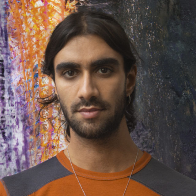 Portrait of young man with orange shirt and shoulder-length black hair, in front of a printed fire background. PC: Yusef Audeh