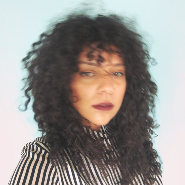 Blurry image of an AfroCaribbean woman with long curly hair wearing a striped black and white button down shirt in front of a blown out turquoise / white background.