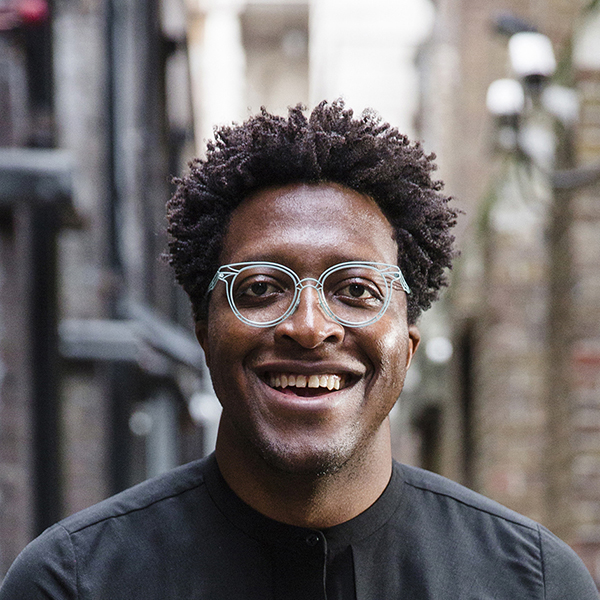 A young African man with an open mouth smile showing teeth, he's wearing a black collar-less shirt and blue glasses which contrast against his dark brown skin. He is standing in an outdoor urban space.