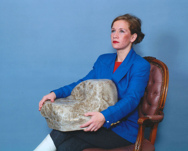 A young woman with fair skin sits in a 1970s Victorian Style chair, photographed in a portrait studio with bright lighting on an all blue background. Her light brown hair is pulled back into a bun and she is wearing heavy foundation and red lipstick. She is wearing a bright blue blazer and she clutches a rock that is almost as large as her, as she looks up.