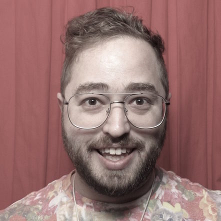 Harris in a photobooth with red backdrop. Harris is white with light brown hair, glasses, and facial hair, and is wearing a t-shirt with a collage of cats on it.