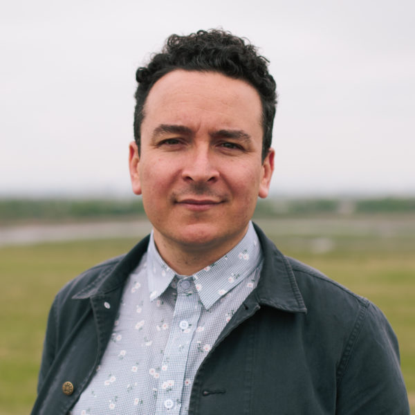 This is a headshot of Dylan Gauthier, an artist and curator, who presents as a white male with curly black hair, a sharp nose, and an uneven chin. He is wearing a black worker's shirt over a flower print white and black button-up shirt. The blurred background hints at a view of Freshkills Park, where the photo was taken. The photo was taken by Natalie Conn.