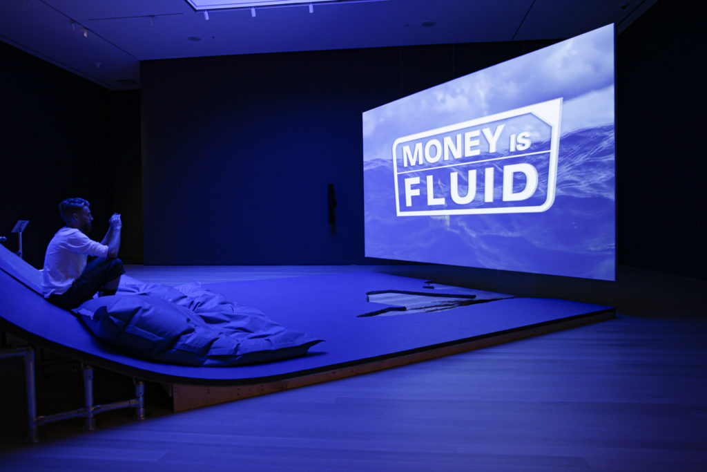 Photo from Hito Steyerl exhibition, lit in blue you see a screen that reads "Money is Fluid_