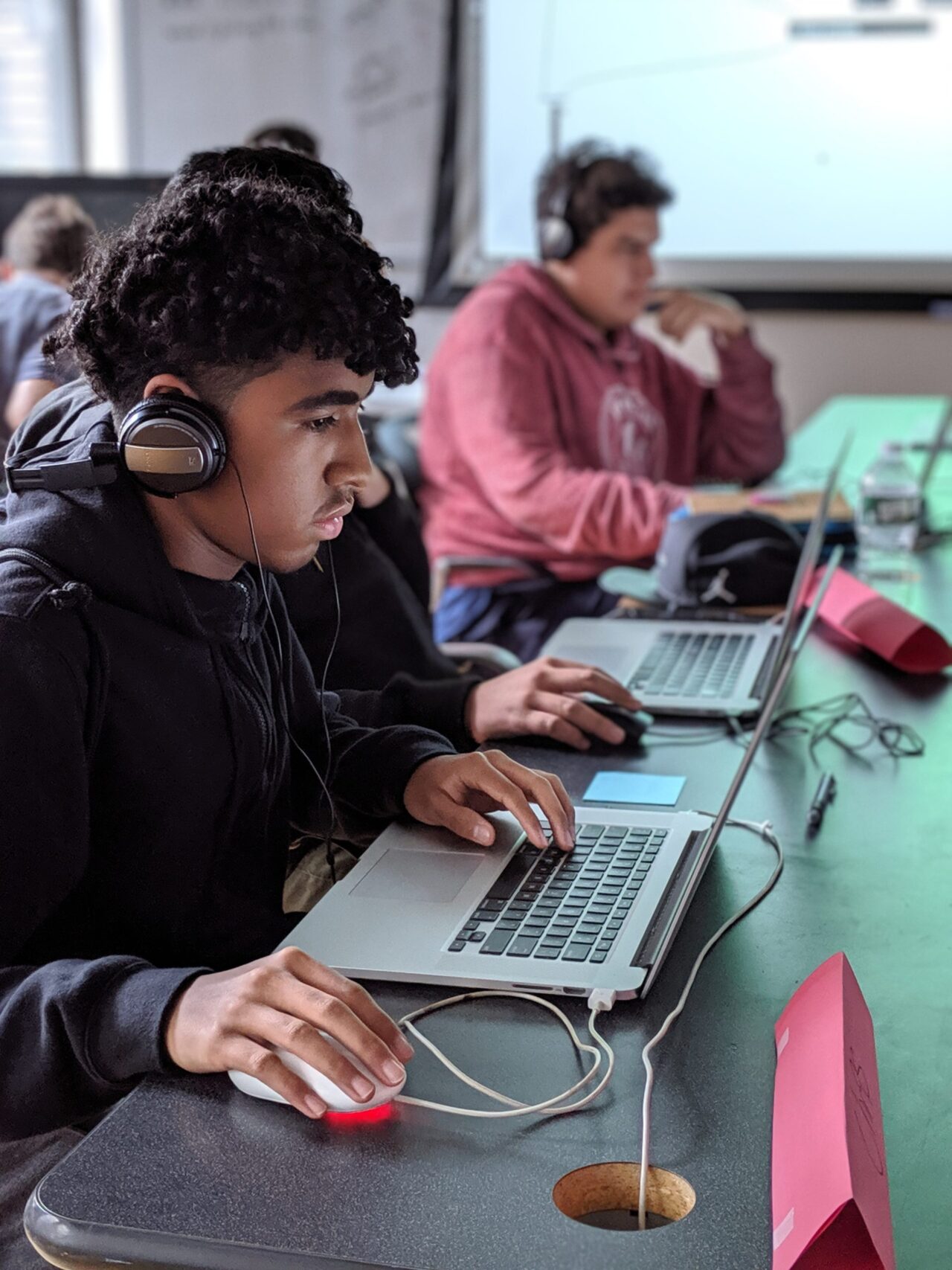 Three DDC 2019 Students working on tan apple laptop computer learning Max, in front of a white screen projector. They focus on their screens in front of them with over the ear headphones on, following the lesson.