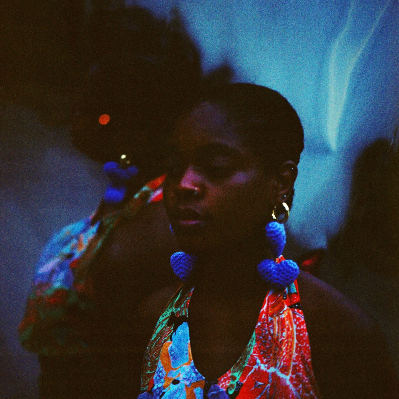 Neta, a deep brown-skinned person, wears a colorful halter top and large lavender heart-shaped crochet earrings as she stands in front of a reflective backdrop that doubles and distorts the image of her profile.