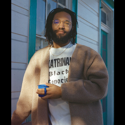 A man with brown skin, medium length dreadlocks wearing gold glasses stands behind a blue shotgun home on his balcony wearing a light brown cardigan with a t-shirt reading "Katrina! Black Genocide" as he holds a blue enamel coffee cup looking at the camera directly with a small smile apparent.