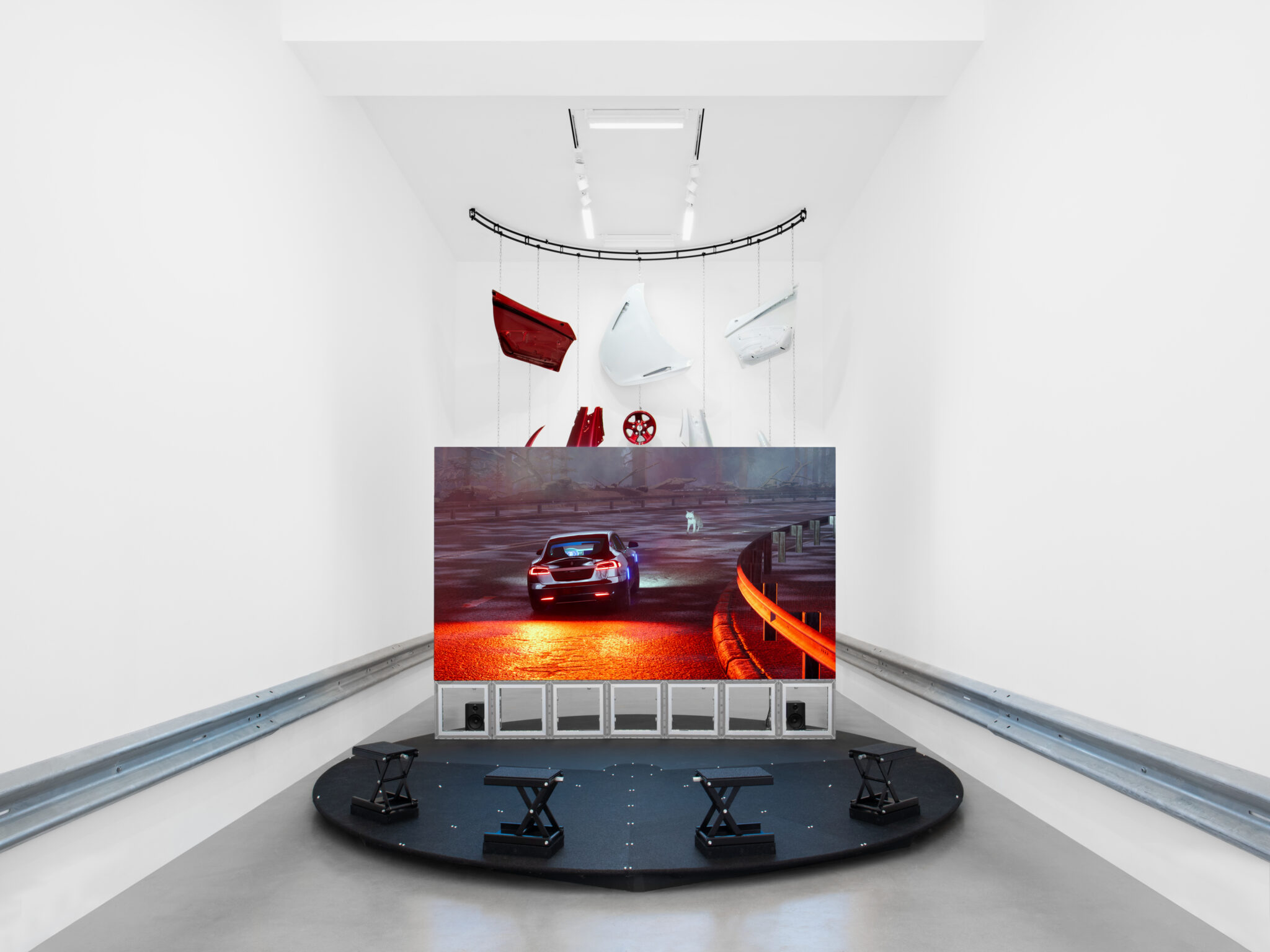 Pictured is a photo of installation, which at its center, the 11-minute film is framed by highway barriers and suspended body panels from damaged supercars, alluding to the aftermath of a road accident. Arrayed around a single wheel, the panels are refinished in candy apple red and sonic blue — colours used in both automotive and electric guitar design, reflecting Lek’s longstanding interest in the relationship between industrial fabrication and music production.