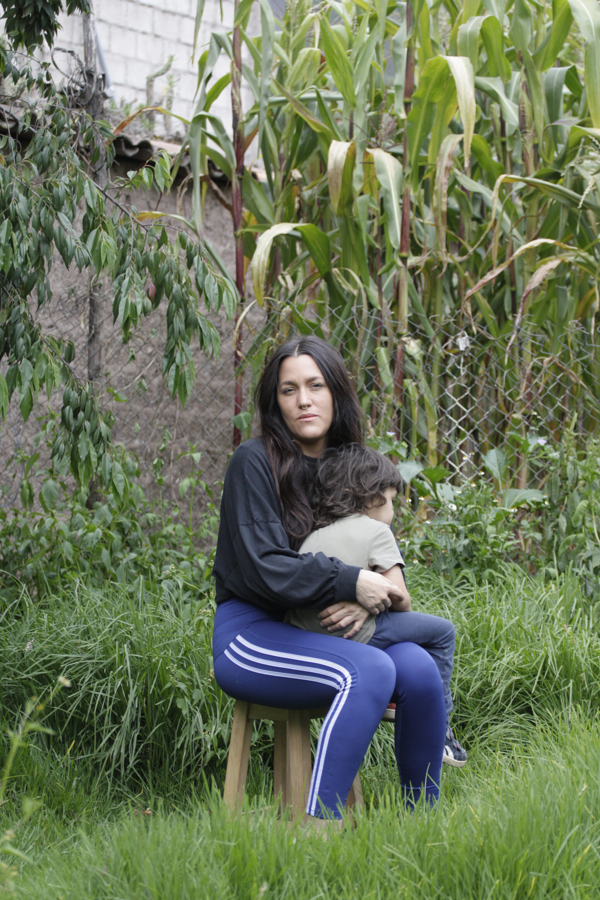 Daniella, a fair-toned woman with long black hair wears a black top and blue sweatpants with white stripes on the side, sits on a wooden stool in a garden, while holding a toddler.