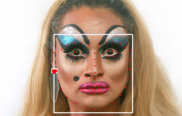 A white person with long, blonde hair, blue eyeshadow and pink lipstick with a white square framing their face with red facial recognition dots