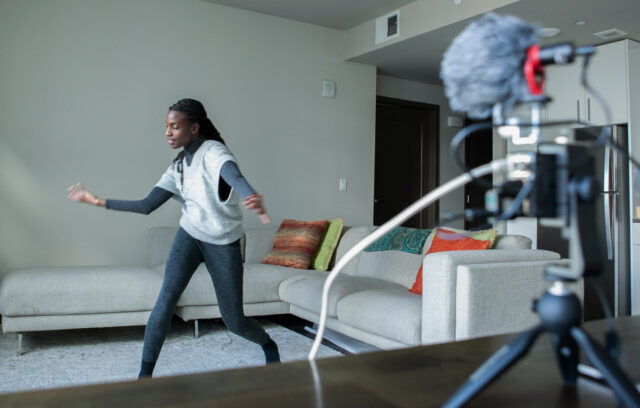 Valencia, a black woman with long black hair in a gray outfit, strikes a dance pose, facing a small, black camera on a short tripod with a gray dead cat.