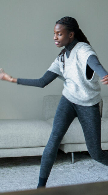 Valencia, a black woman with long black hair in a gray outfit, strikes a dance pose, facing a small, black camera on a short tripod with a gray dead cat.