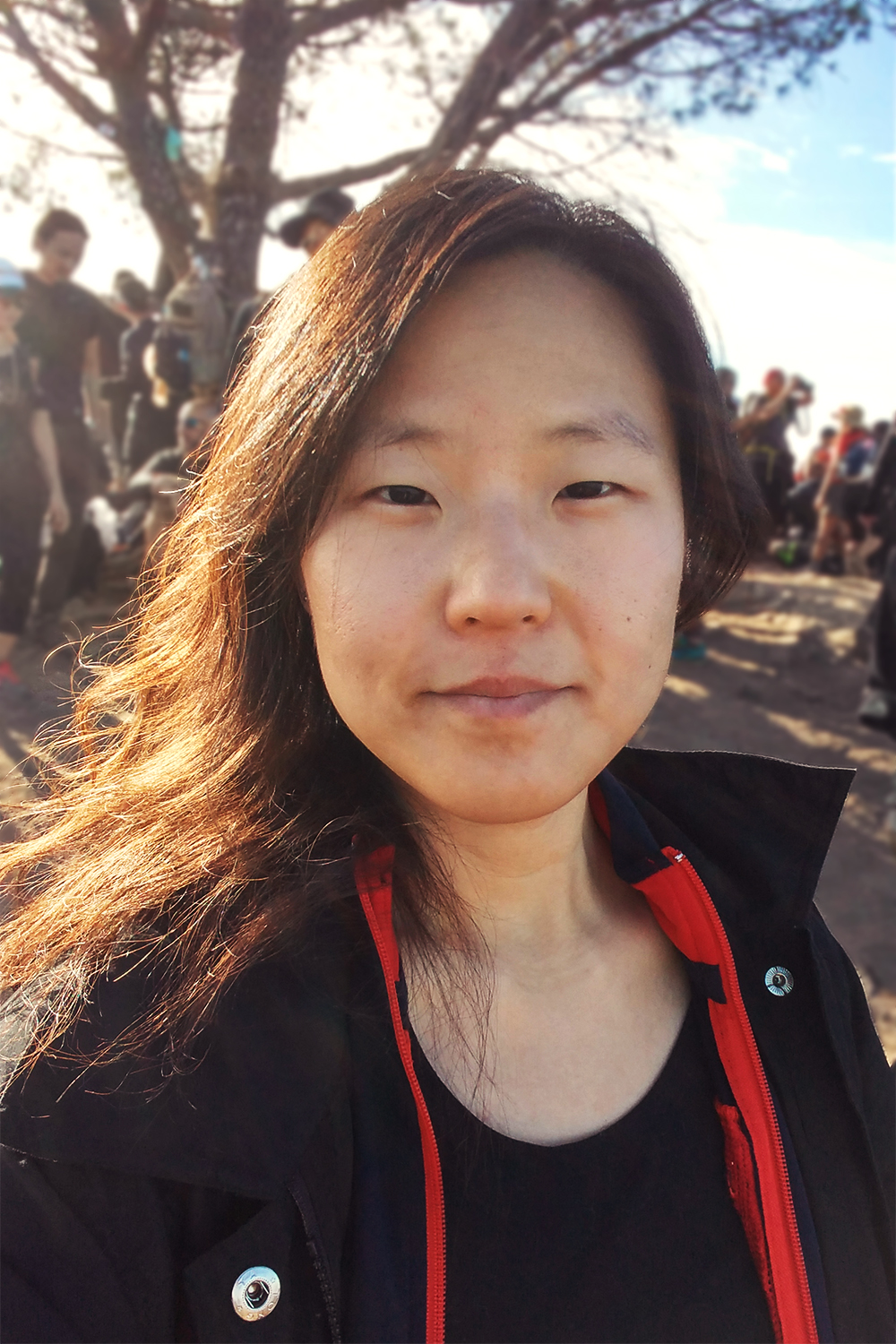 An image, taken from the chest up, of an adult Korean woman in casual clothing, with a slight smile, and with long untidy black hair swept to her right shoulder. She is standing outdoors in daylight, and warm sunshine lights up the right side of her face, mostly her hair, which reflects yellow and brown. In the blurred background are a number of people further away standing around, indicating that she was part of a group hike activity.