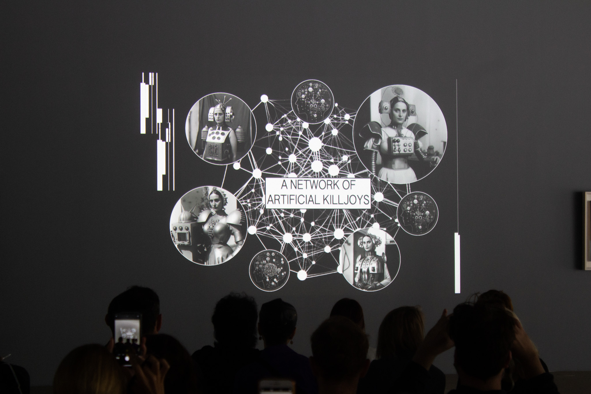 Pictured is a still from a recorded performance. Projected onto a dark grey/black wall, in white/light greyscale imagery is “A Network of Artificial Killjoys,” and a graphically intricate representation of a network, multi-nodes all connected in a hive-like structure.