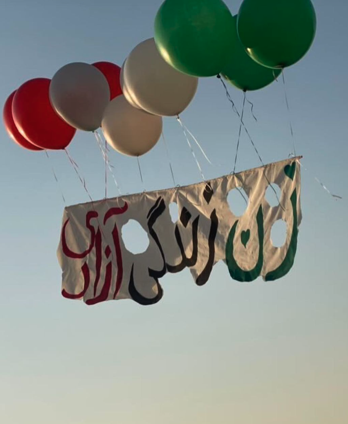 An archived image taken from an anonymous social media source. A photo aimed up at the near-dusk light blue to yellow-orange sky, are balloons in Red, White, and Green, holding a banner that says Zan Zendegi Azadi [Women, Life, Freedom] in farsi.