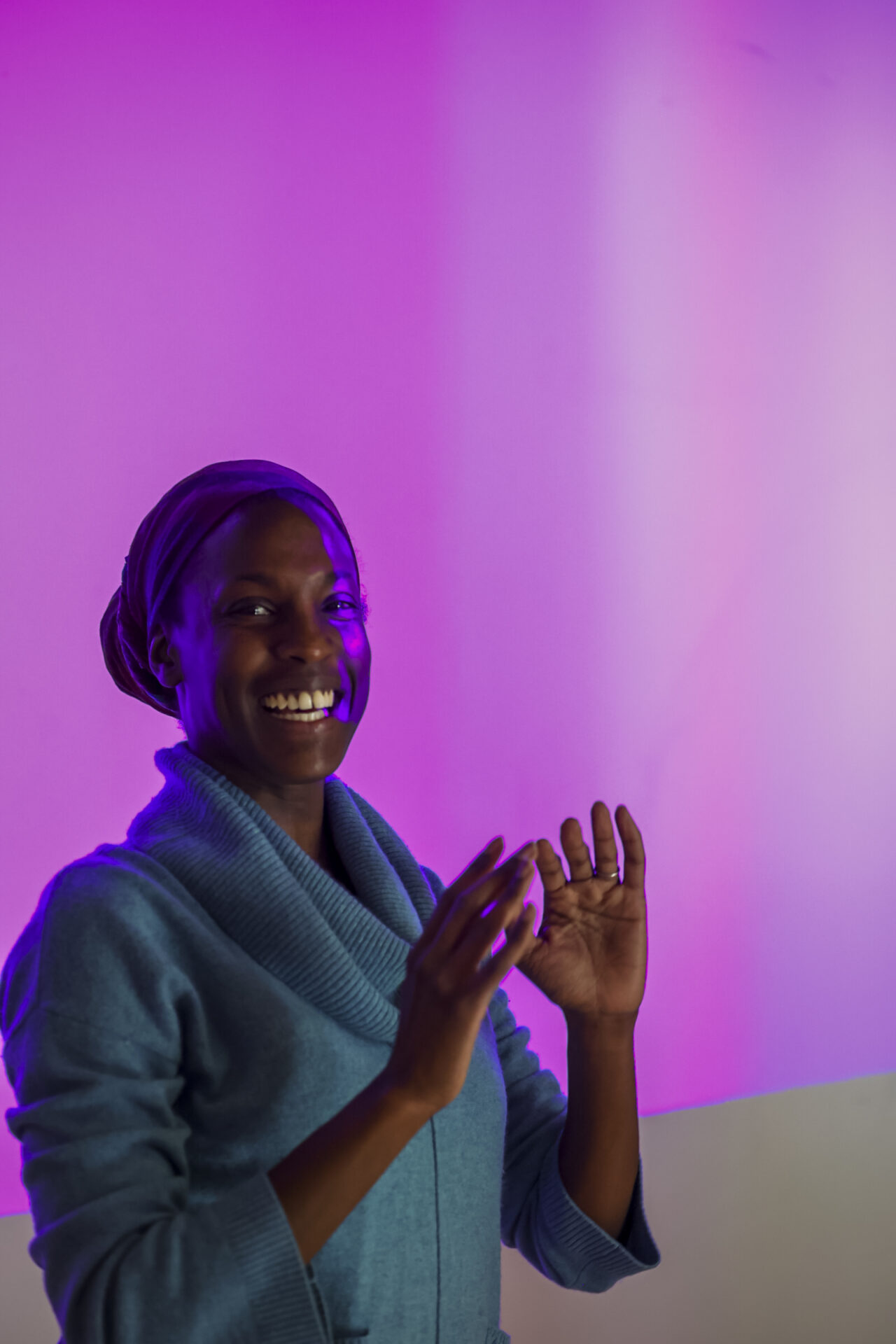 Pictured is Valencia James, a Black dancer wearing a teal baggy-collared sweater, who is smiling, baring her teeth and facing directly at the camera. She stands with her arms bent in front of her, palms facing up as she demonstrates her AI and dance project to a room of participants, not pictured. At this moment, a video projection hits the white wall behind her and a portion of her face, painting nearly the entire image with a strong hue of Byzantine, Hex code #AC42A8, rich tone of medium purple nearing Magenta.