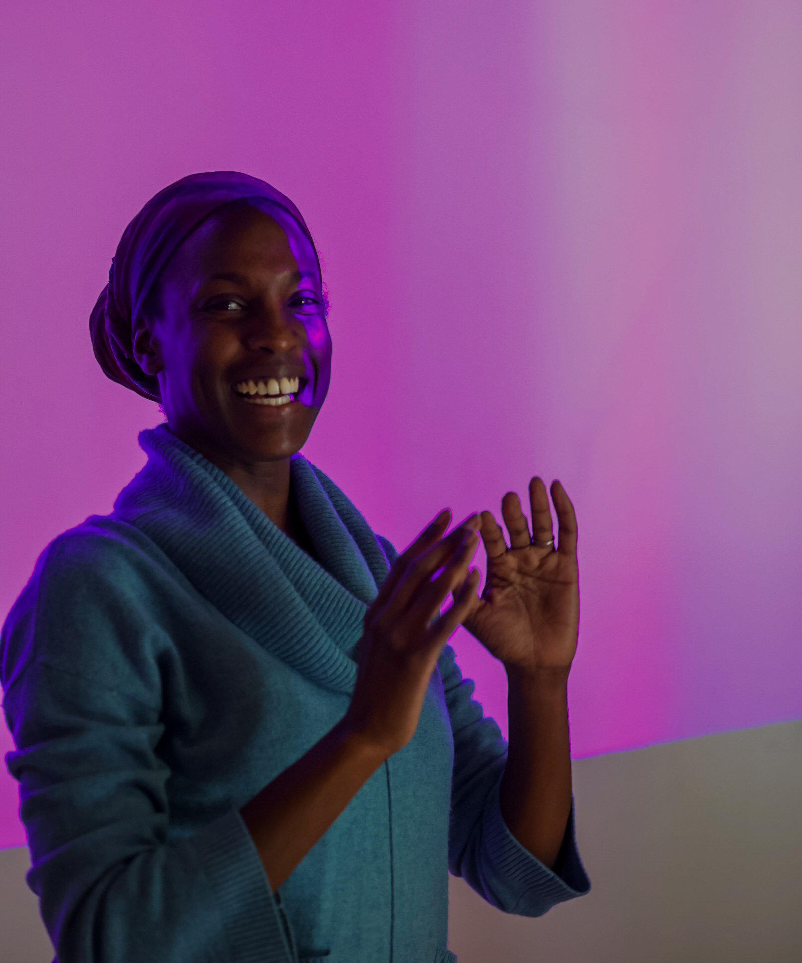 Pictured is Valencia James, a Black dancer wearing a teal baggy-collared sweater, who is smiling, baring her teeth and facing directly at the camera. She stands with her arms bent in front of her, palms facing up as she demonstrates her AI and dance project to a room of participants, not pictured. At this moment, a video projection hits the white wall behind her and a portion of her face, painting nearly the entire image with a strong hue of Byzantine, Hex code #AC42A8, rich tone of medium purple nearing Magenta.