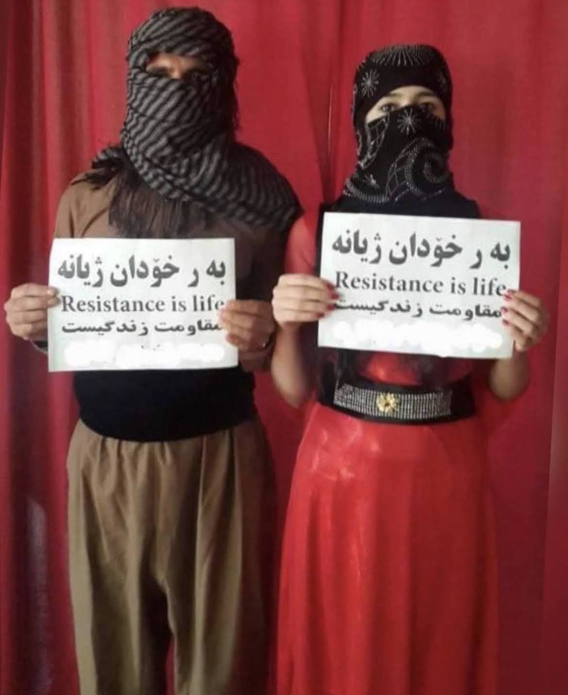 An archived image taken from an anonymous social media source. In the photo are two anonymous women, with faces covered by dark grey/black scarves, holding up printed signed that say “Resistance is life” in both English, Arabic, and Farsi. They stand in front of a deep red curtain.