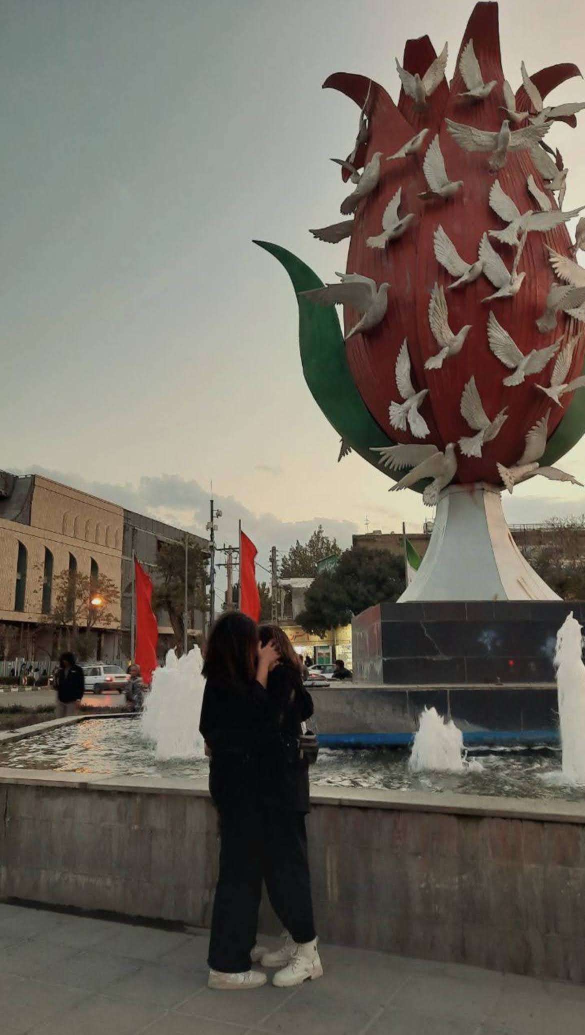 An archived image taken from an anonymous social media source. In the photo are two anonymous women, hiding each other's faces, facing each other, all dressed in black with white shoes. They are in the midst of an intimate embrace and kiss out in public, in what seems to be a common square. They are standing right by a large water fountain that has a red flower covered in white doves as a centerpiece. Red banners and people in the distant background can be seen. This is a photo taken to demonstrate queer resistance.