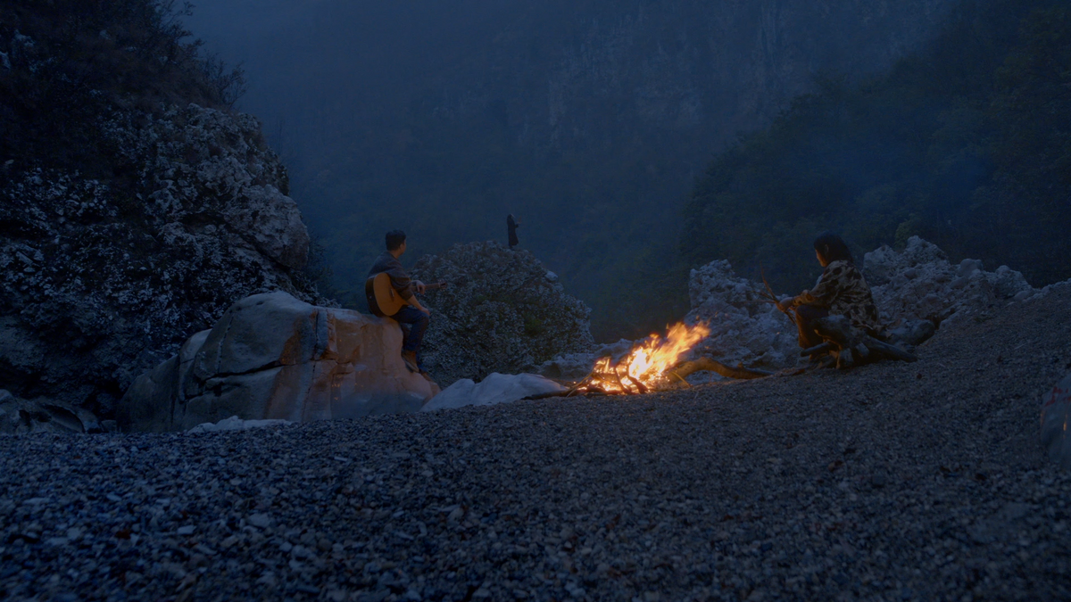 Two people are sitting out in the edge of the woods, around a fire. The man is seated on a large smooth rock, holding a guitar on his lap, watching over the distance, while the woman looks into the fire.