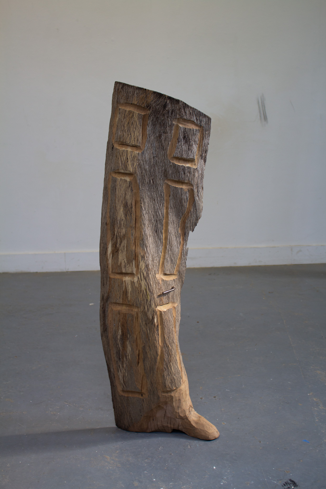 A photograph of a carved and sanded weathered grey cypress-wood sculpture made by Ryan Christopher Clarke. The Cypress lumber has been shapened into a thin, tilting, squeezed door with scrap metal used as a handle. Cypress wood is said to be “extremely durable, light, soft, straight grained with a soft amber or reddish hue and easily workable,...insect resistant, and very stable in contact with soil or water, tending to resist all forms of rot…can naturally age to a weathered gray” 2001 pamphlet by McReynolds Architects, The Menil Collection – Exterior Wall Renovations.