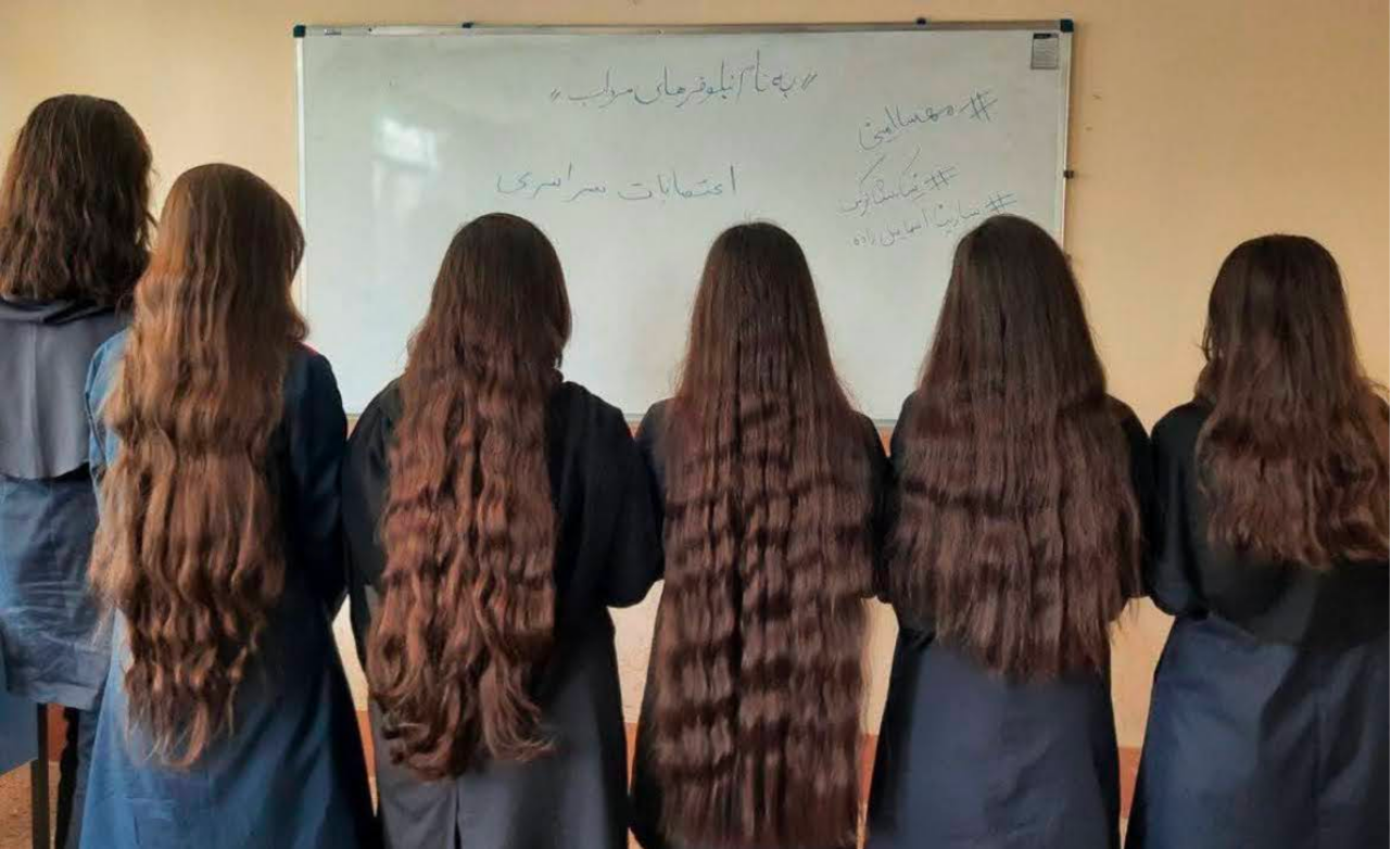 An archived image taken from an anonymous social media source. The image features 6 Iranian girls, in a classroom, with their hair let loose, all of them with long dark brown wavy hair, sans Hijab, in protest against the Hijab laws in Iran. The anonymous girls are facing away from the camera, facing a glossy white board.