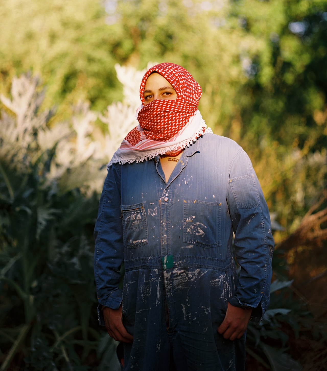 A photograph of Zeina Baltagi with pale brown skin, brown eyes and eyebrows standing in natural foliage. She is wearing blue Lee Can't Bust Em 1940s Union cover-alls, tan work boots, and a red and white patterned keffiyeh scarf around her neck which covers her nose and mouth. Her thumbs rest in her pockets.