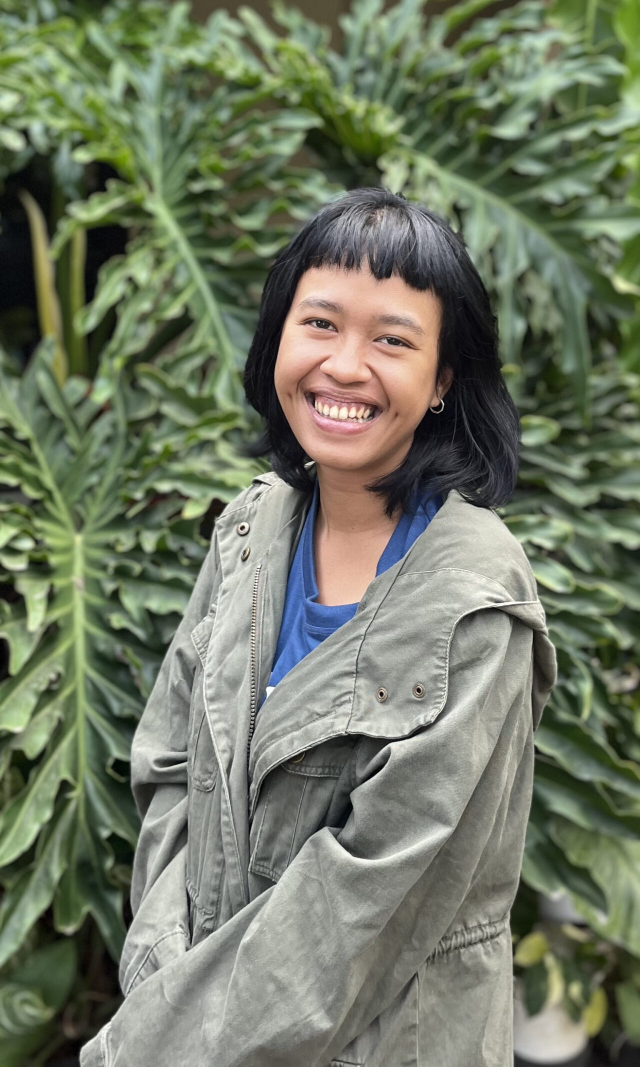 A Photo of the artist Syaura, an Indonesian woman with shoulder-length, straight, black hair. She smiles from cheek-to-cheek, baring her teeth. She stands in front of green tropical foliage.