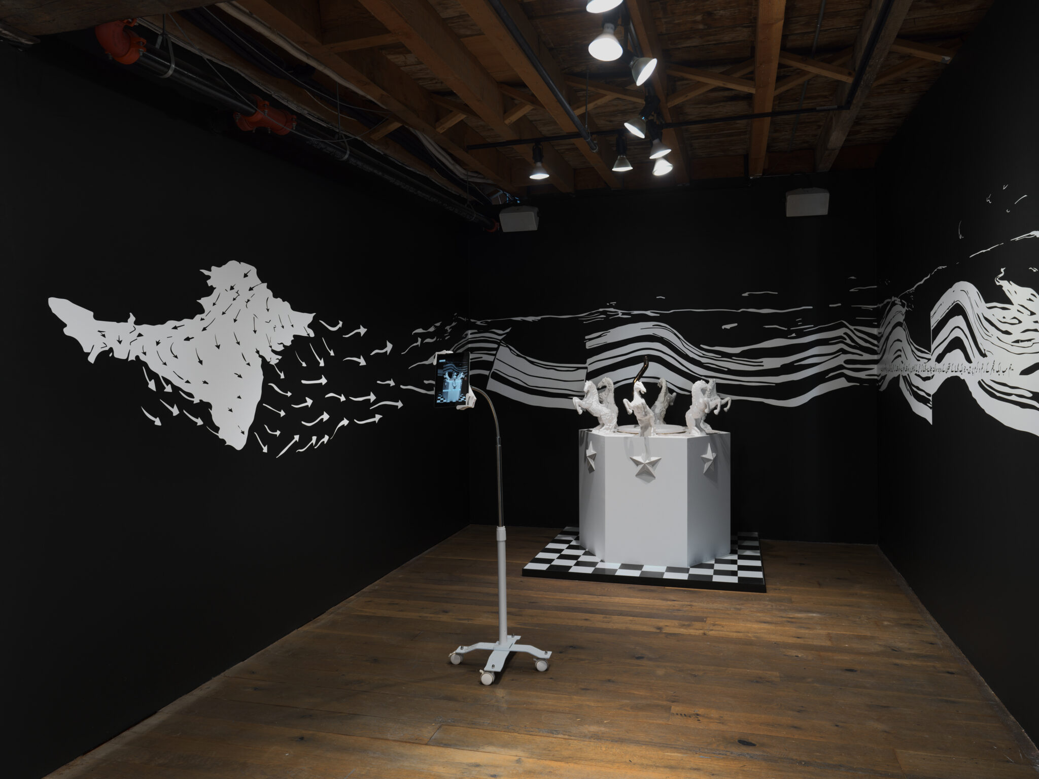 The image at the bottom: In a slightly more dimly lit room side of the room on the right as your enter, with black walls and a white vinyl wall. There is a white plinth on a black-and-white checkered platform, with ceramic horses lined around the perimeter at the top of the plinth. As each side of the hexagonal columns are white ceramic stars. Facing this sculpture is a screen with an AR animation display that activates the sculpture, what you now see is a fountain with flowing water, the water made from the textures of scanned plastic bags that Umber found on the ground, littered material from the developments in Lahore.