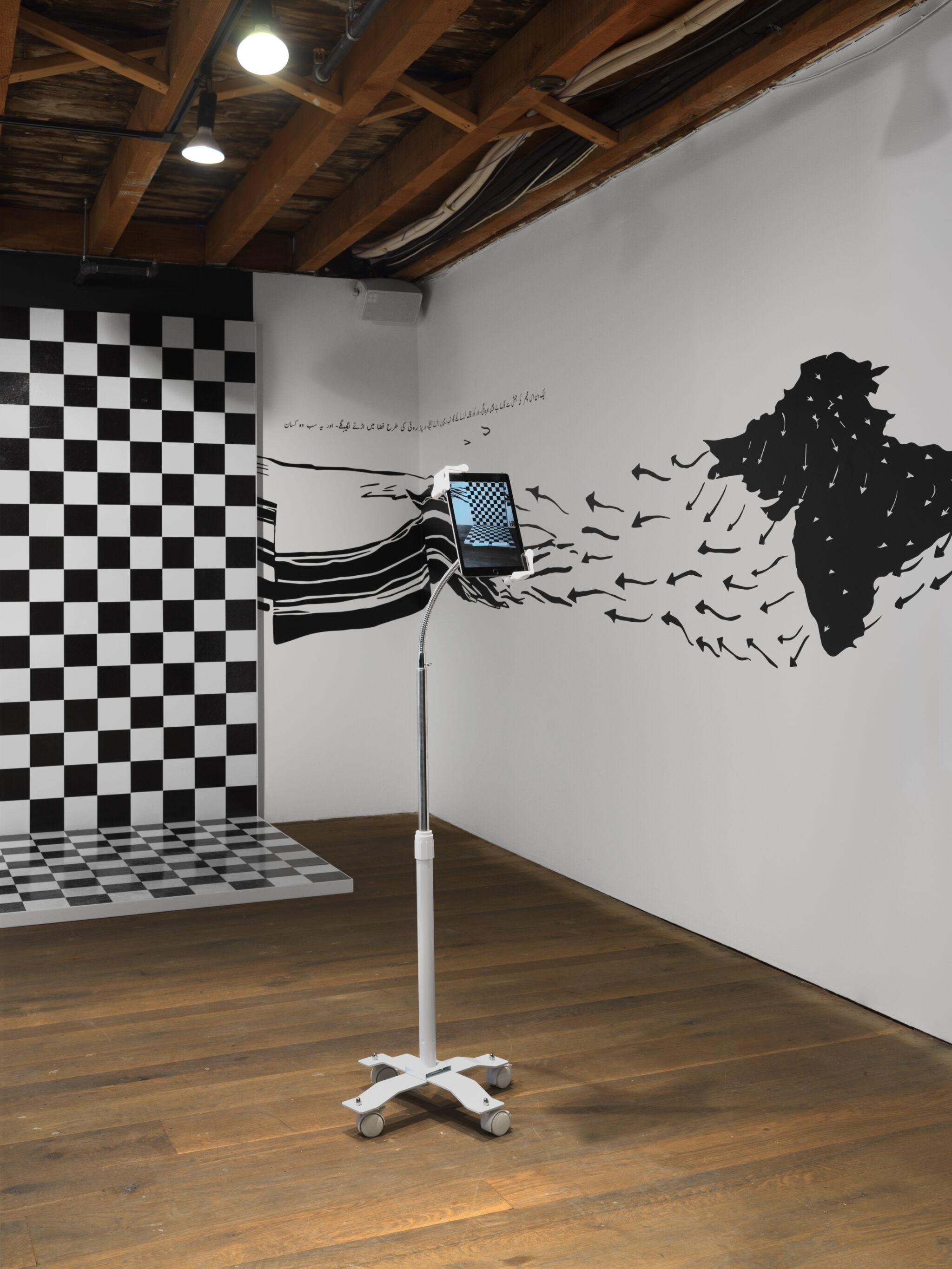 The top left: There is a black-and-white checkered pattern on the wall and floor in the middle of the room, a platform where people can take photos. Facing that is a monitor on a stand display that takes photos, on the screen are repeated AR animations that play. On the wall, flowing towards the center are Black vinyl, the borders of Pakistan, India, and Bangladesh merged as one Black vinyl sticker. Arrows forming into birds point all out towards the checkered platform from the adjacent walls. On the white walls, you can also see Urdu text undulating with the Black melting waves.