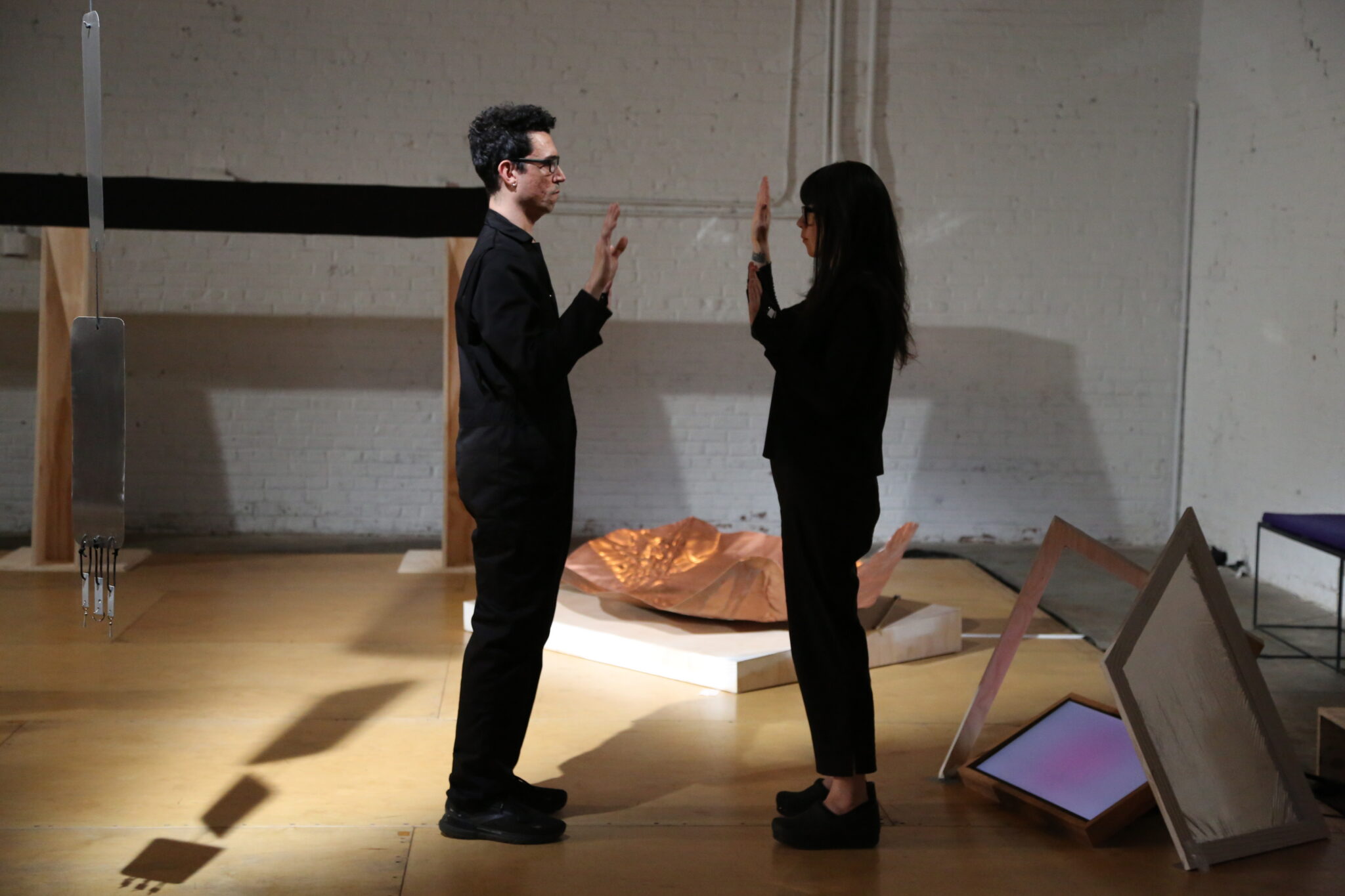 Two people stand facing each other in a dimly lit art gallery. They raise their hands, mirroring each other. Abstract sculptures surround them, creating a contemplative atmosphere.
