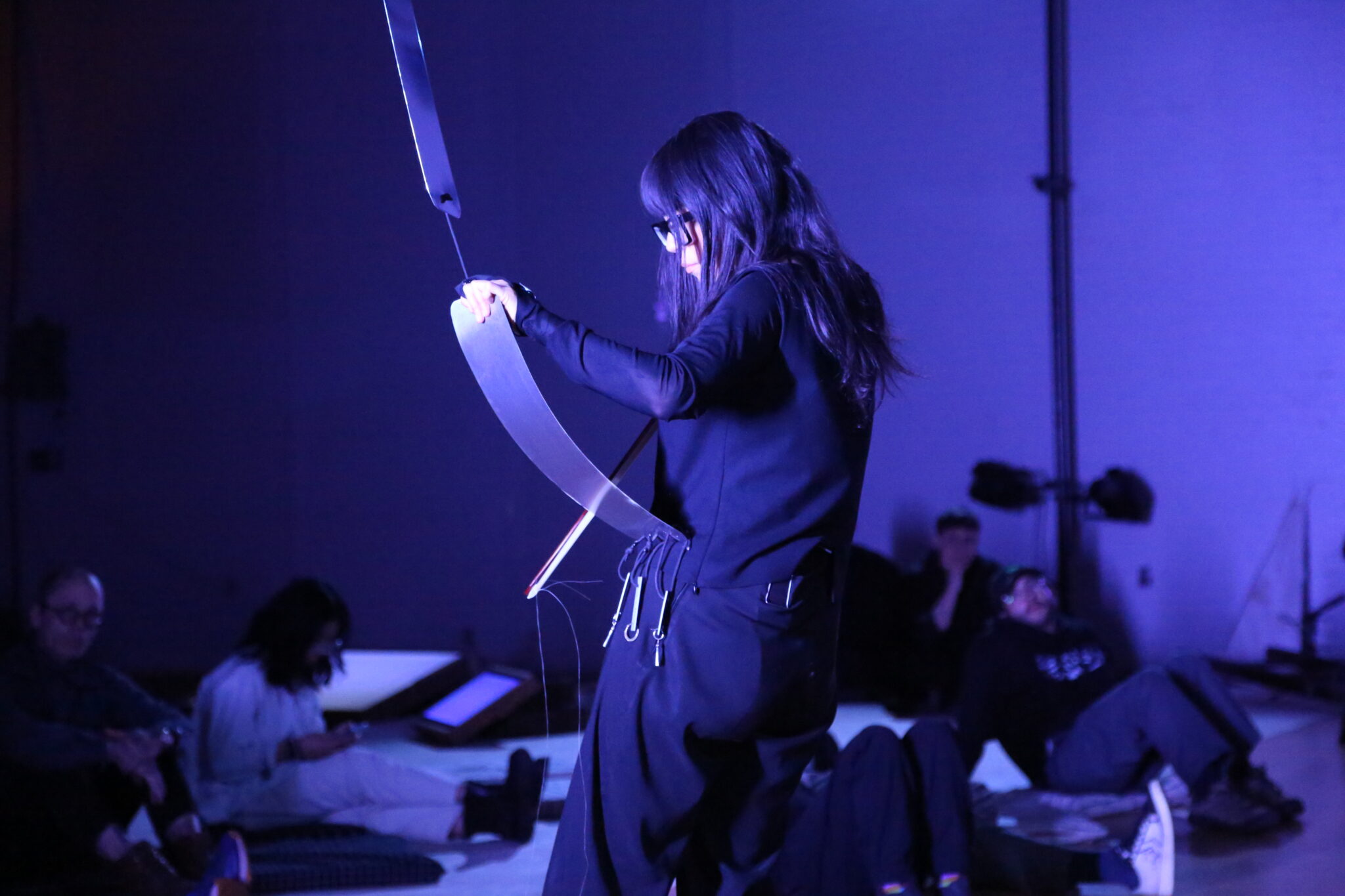 Person dressed in black performs with a metal strip, using a violin bow to create sounds on a bent thin sheet of metal, that's connected to a series of metal hanging from the ceiling. The space is under blue lighting, creating a contemplative atmosphere. Seated audience watches intently.