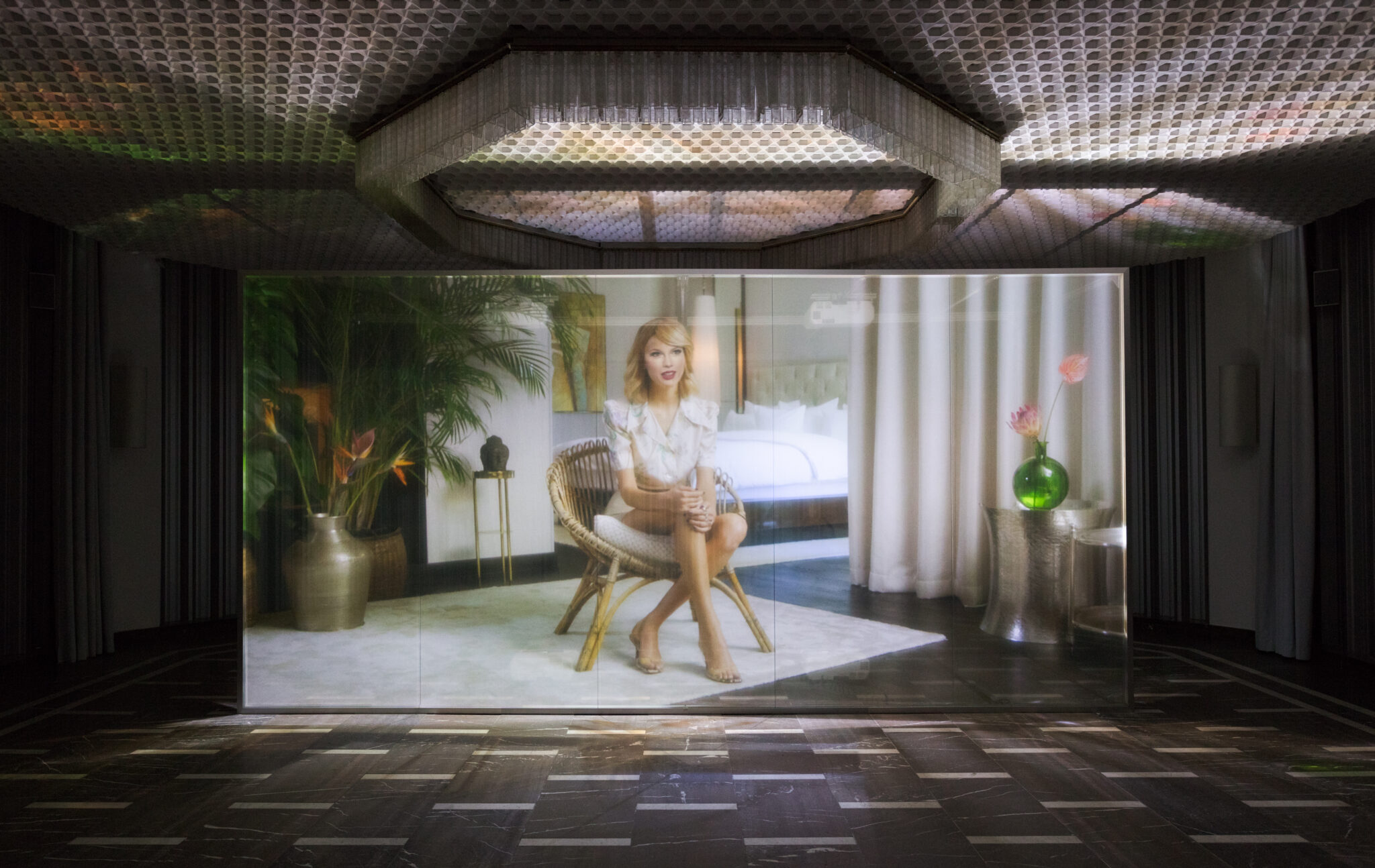 A deepfake video still of singer Taylor Swift, projected on a large rectangular screen in a dimly lit room. The deepfake Taylor Swift sits on a woven rattan chair, wearing a white short-sleeved dress, sitting crossed leg with hands on her right knee.
