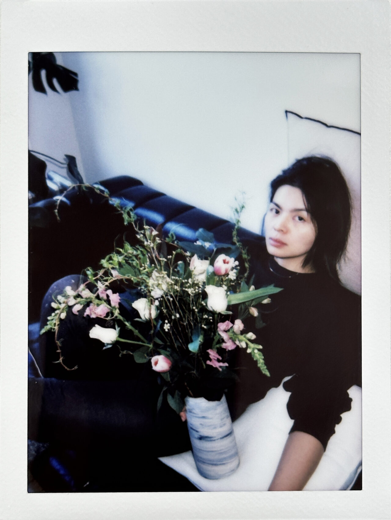 A lo-fi polaroid photo of artist Doreen Chan, sitting on a black leather sofa behind a bouquet of light pink and white flowers.
