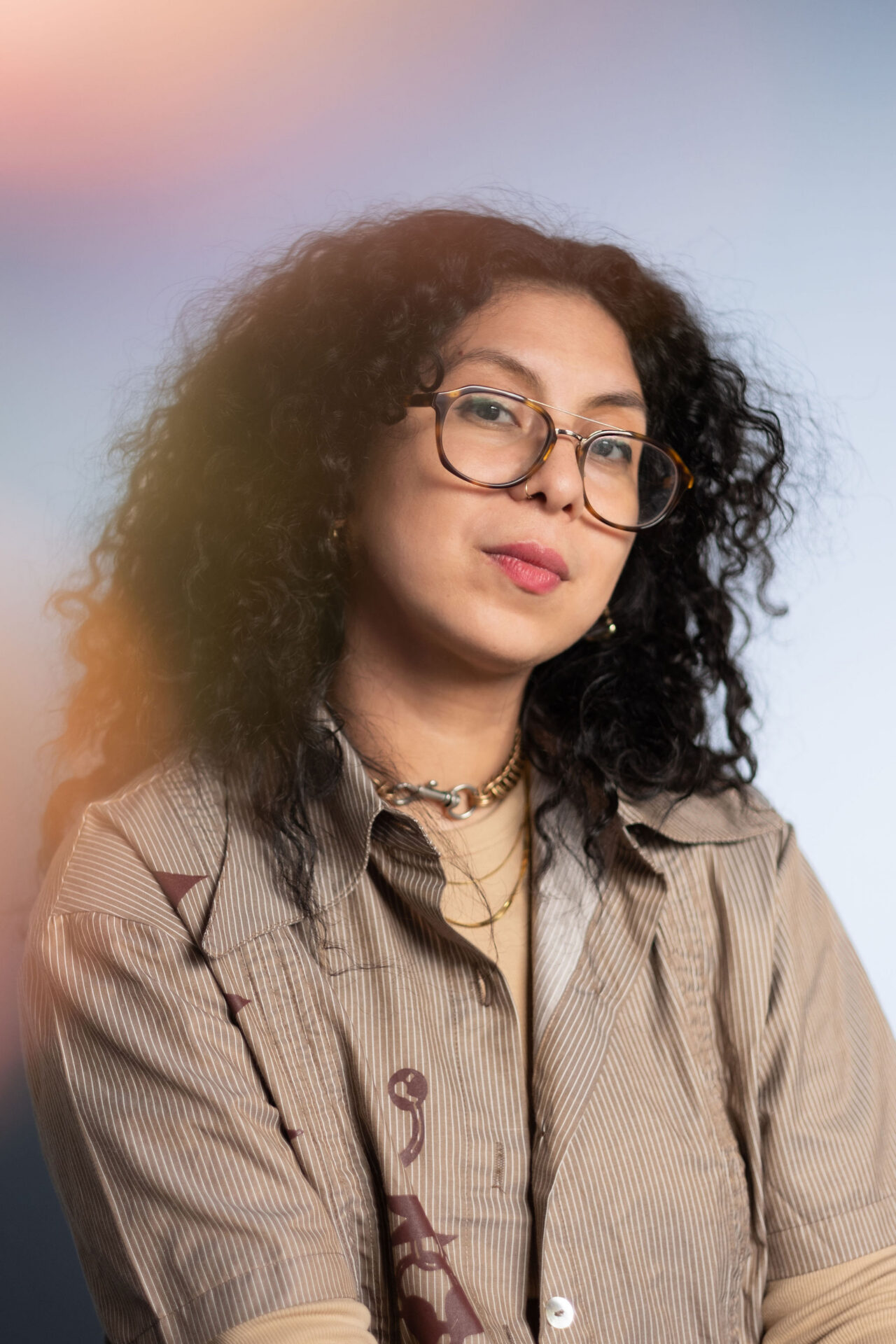 Pictured is artist, dre jácome, a brown femme with big black curly hair with gold hoops and nose rings smiles wearing a light cool brown shirt.