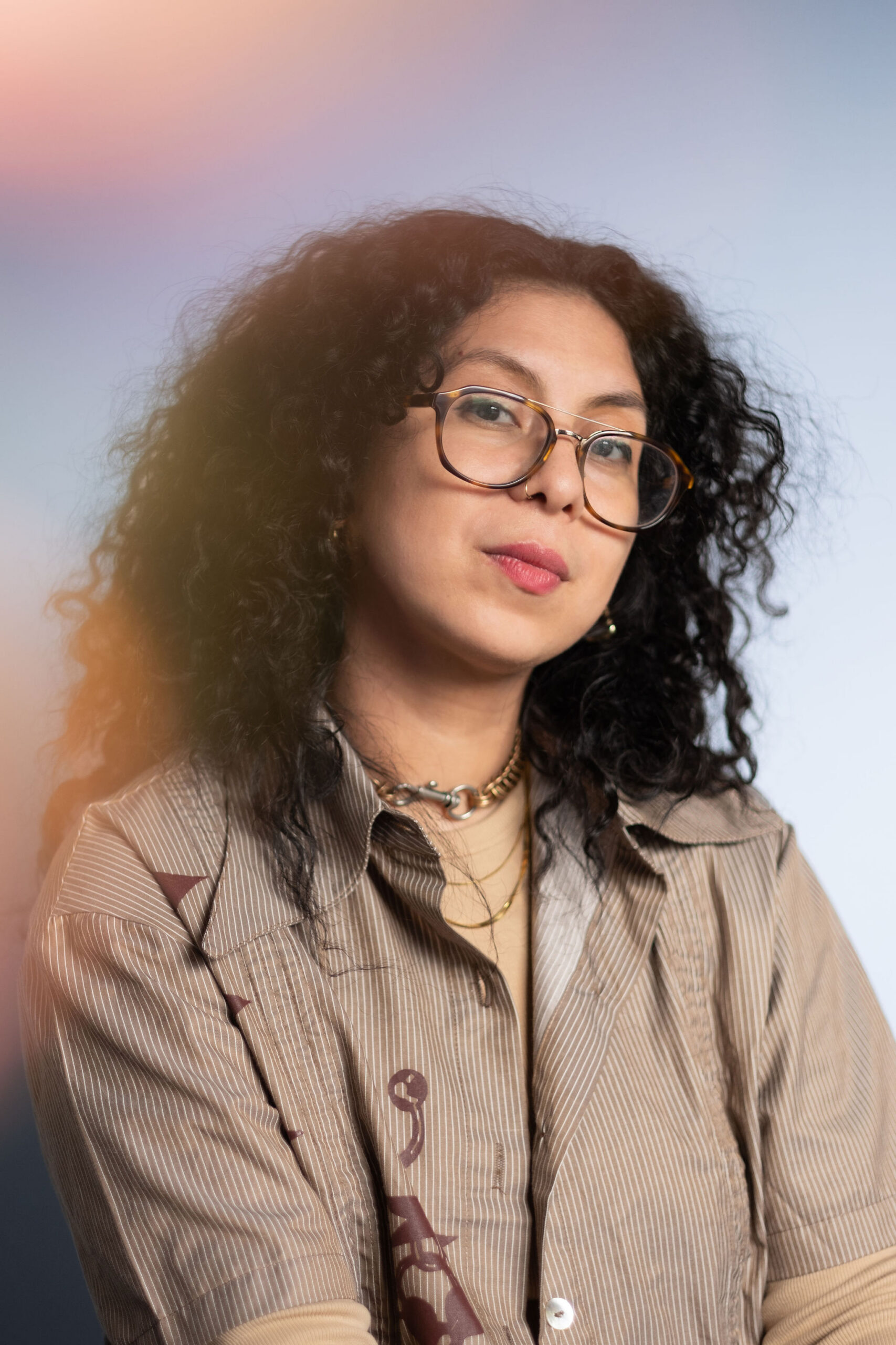 Pictured is artist, dre jácome, a brown femme with big black curly hair with gold hoops and nose rings smiles wearing a light cool brown shirt.