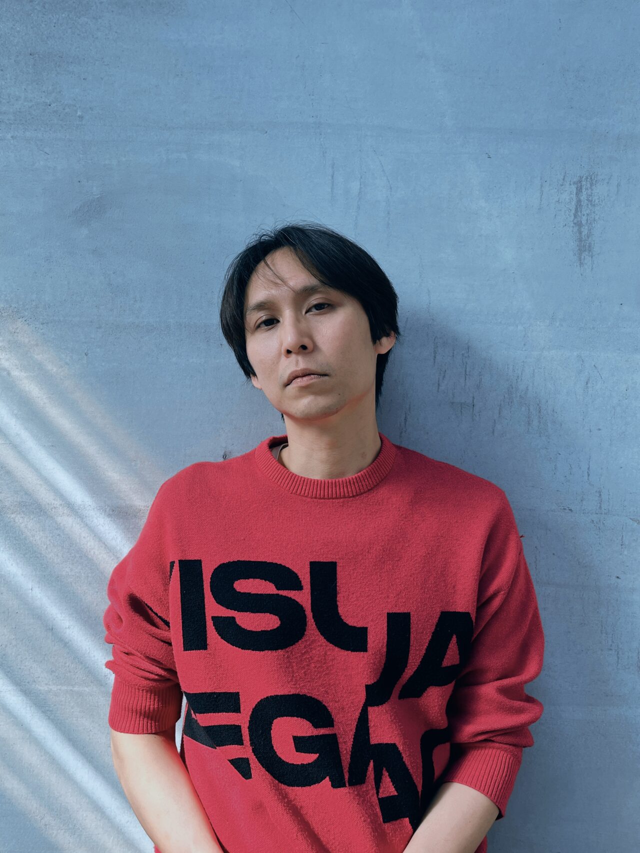 Pictured is the artist Su Hui-Yu, a Taiwanese man with dark straight short hair. He wears a bright carmine pink sweater and rests against a light blue wall in this portrait.