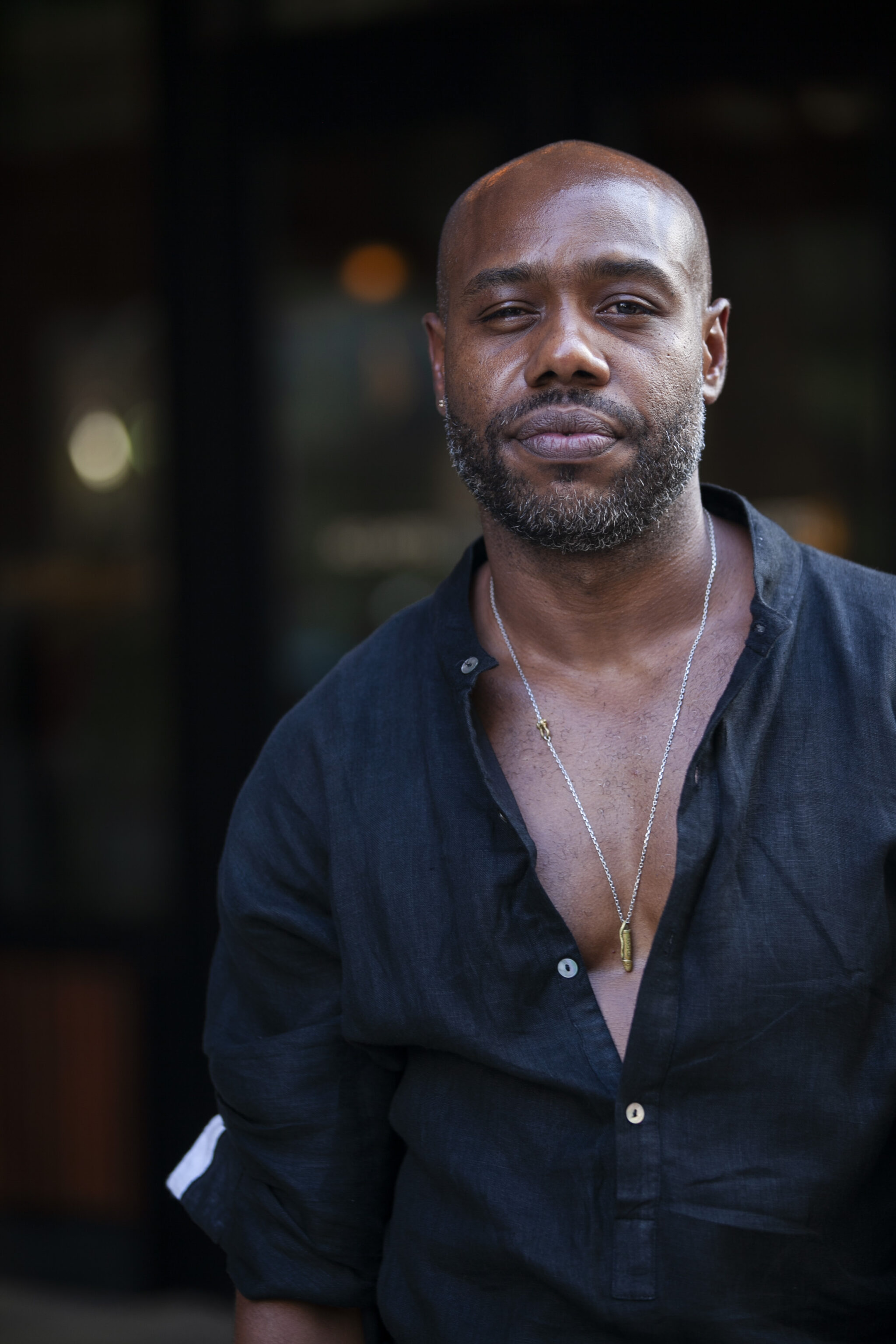 Pictured is artist, Marton Robinson. A dark-skinned bald Black man with a five o clock shadow. He wears a relax fit, black linen button up shirt, slightly unbuttoned to reveal his necklace.