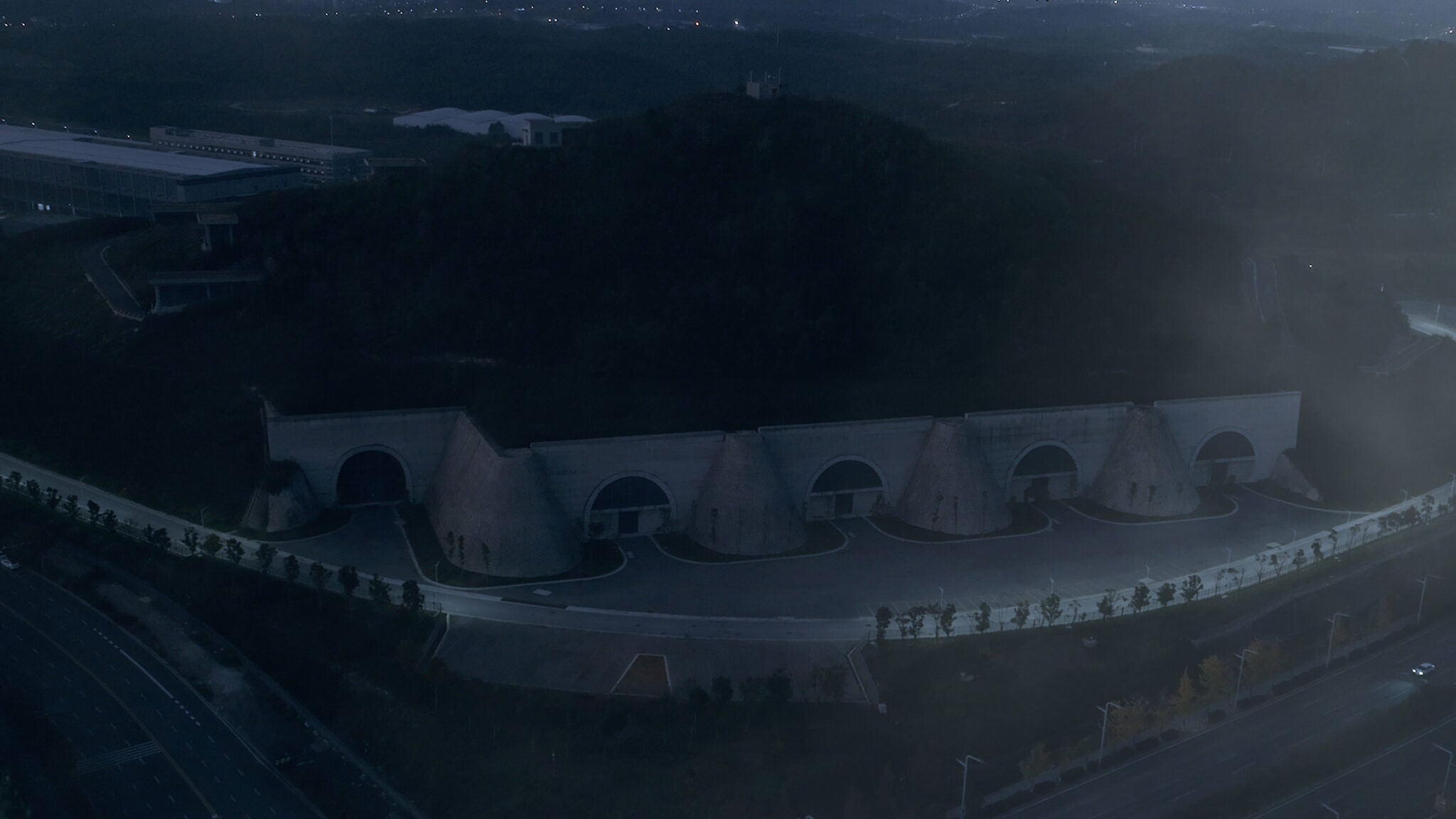 A Large concrete structure jutting out from a tree-covered hill. The street surrounding this facility is neatly lined with trees. Nighttime.