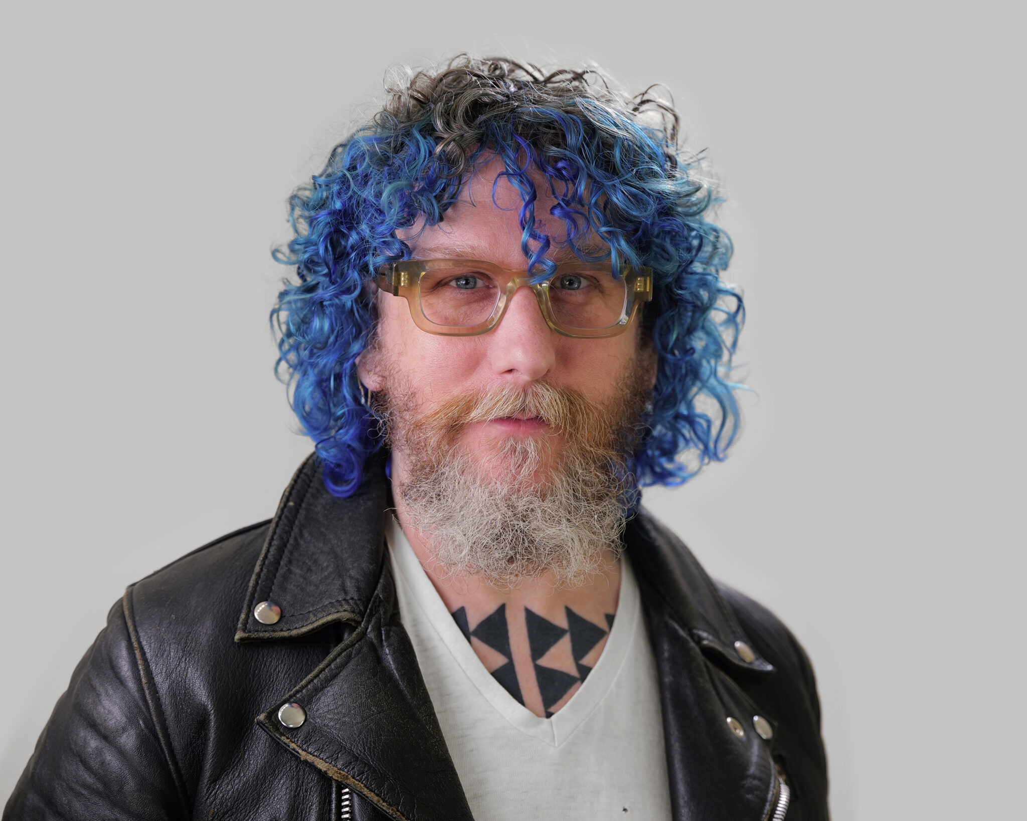 A headshot of Chris, a white person, with blue medium curly hair and white facial hair, wearing glasses, a black leather jacket, gray top and black triangular tattoos on their chest.