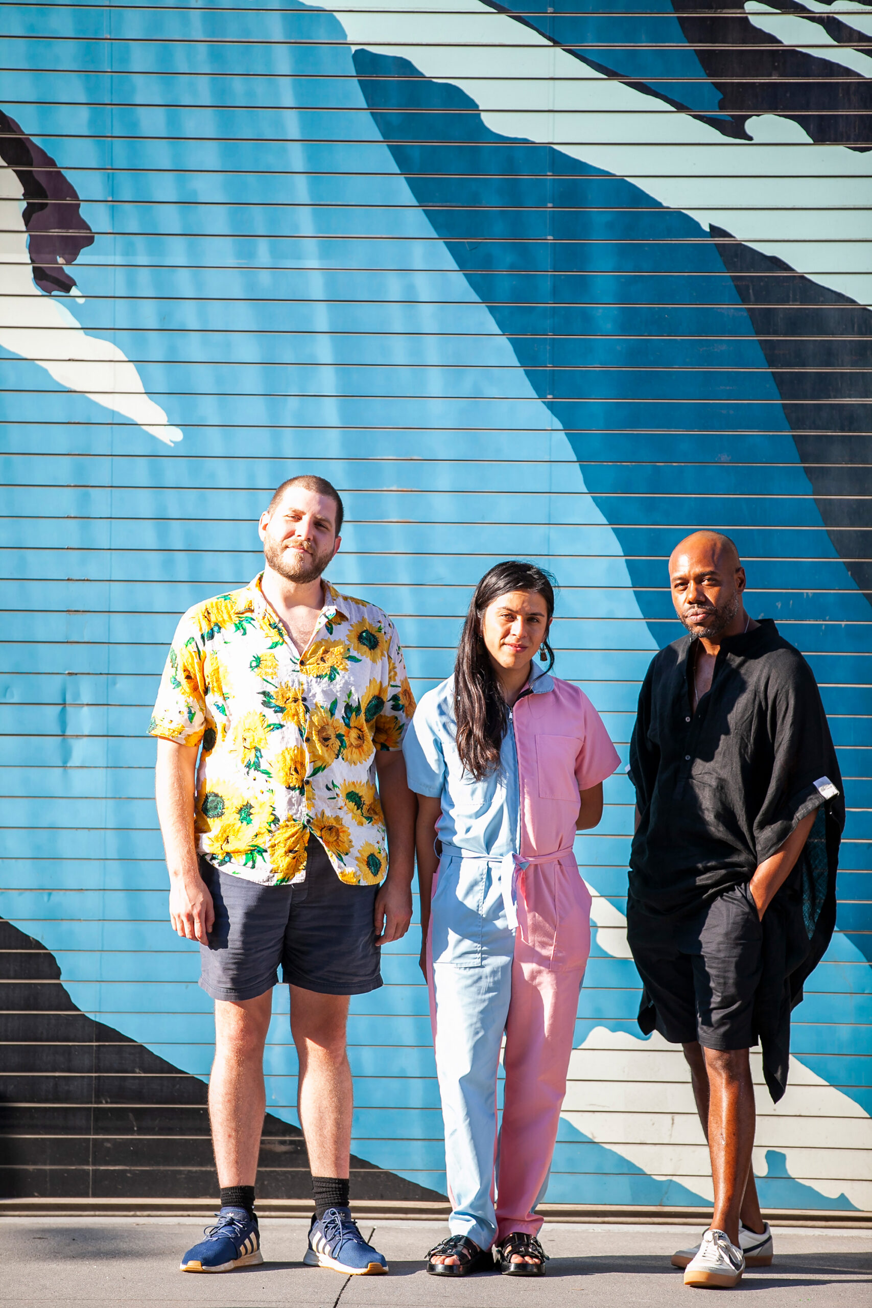 Roo a light-toned male with a flowery top and gray shorts, Seba, a medium-toned person in a pink and gray jumpsuit and Marton, a black man in all black shorts set, stand in front of a blue and black mural.