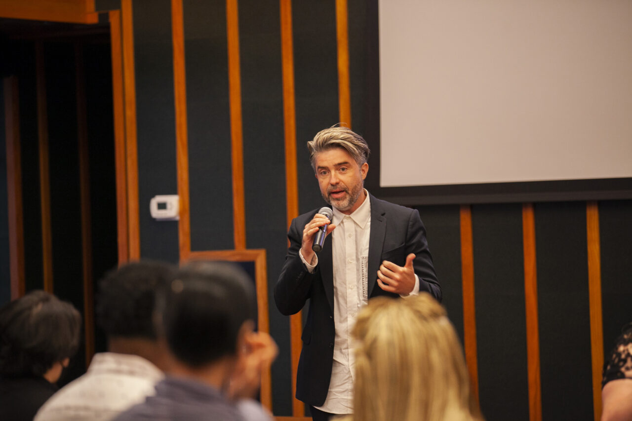 Pictured is Roddy Schrock, the executive director, speaking in public, in front of an audience at the ace hotel. He has salt and pepper hair, is a fair-skinned man in his 40s who wears a black relaxed fit blazer and white button up.