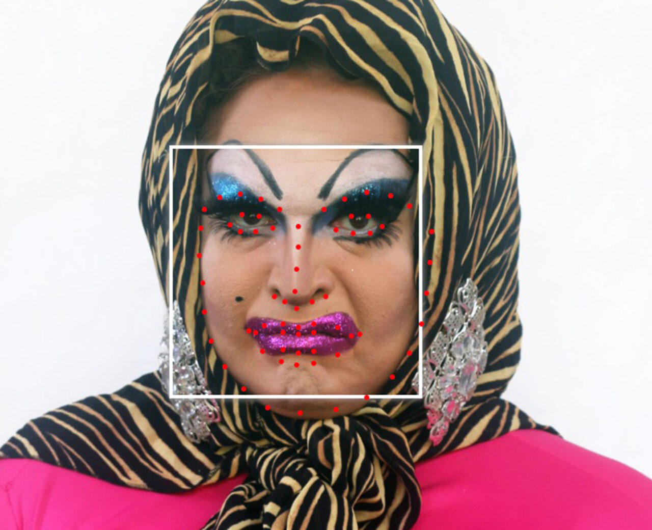 A white person with a black and yellow head scarf, blue eyeshadow and pink lipstick with a white square framing their face with red facial recognition dots.