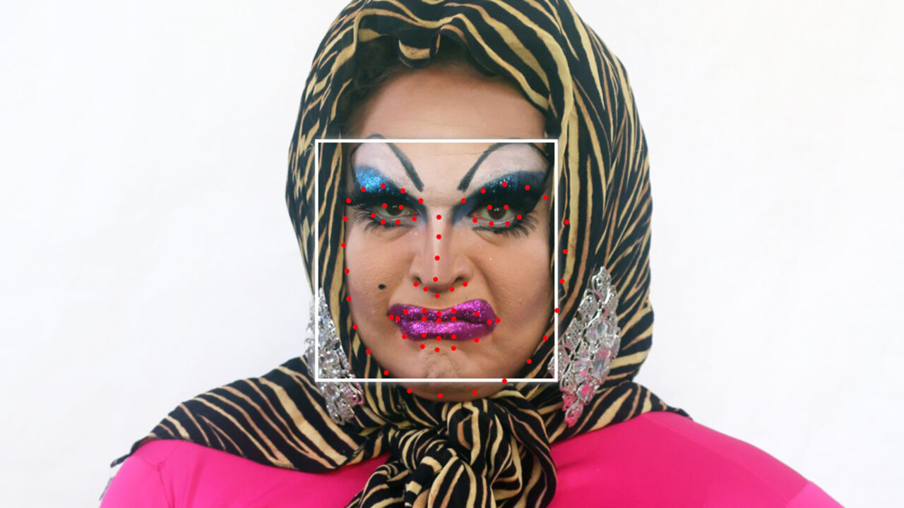 A white person with a black and yellow head scarf, blue eyeshadow and pink lipstick with a white square framing their face with red facial recognition dots.