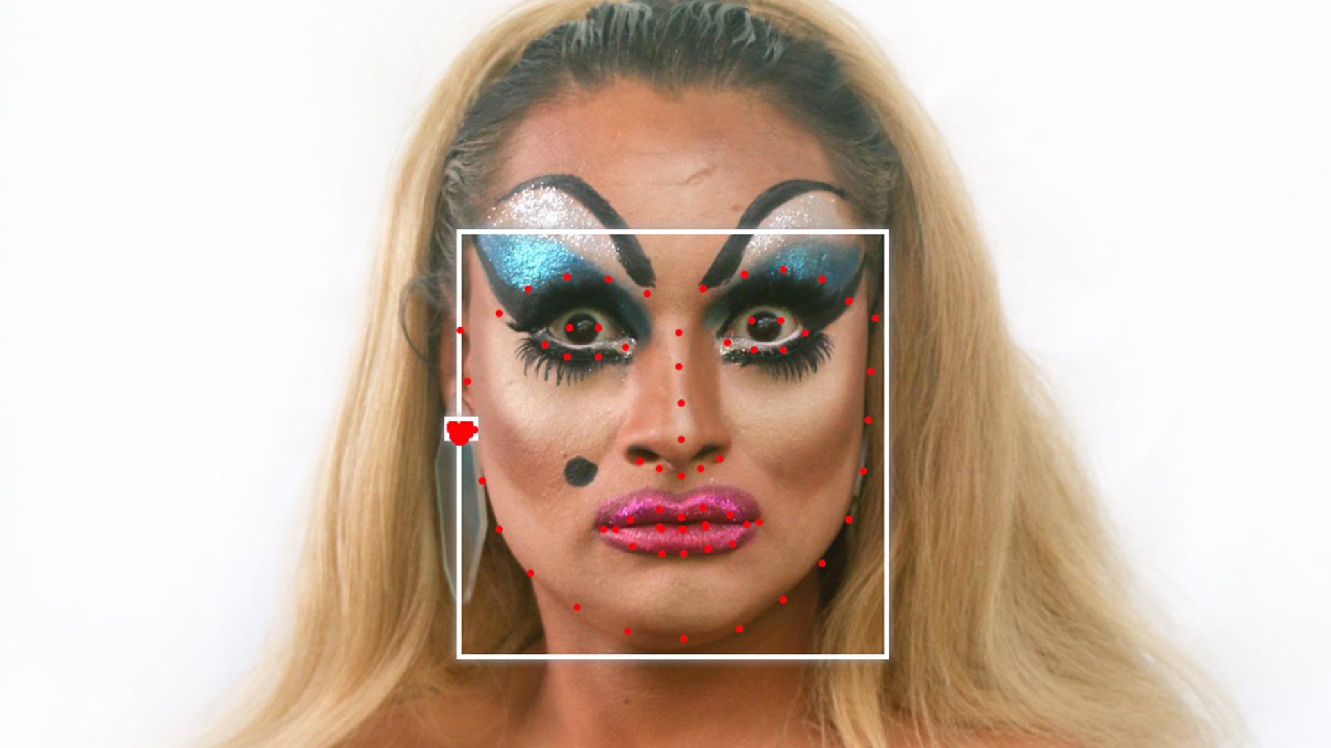 A white person with long, blonde hair, blue eyeshadow and pink lipstick with a white square framing their face with red facial recognition dots.