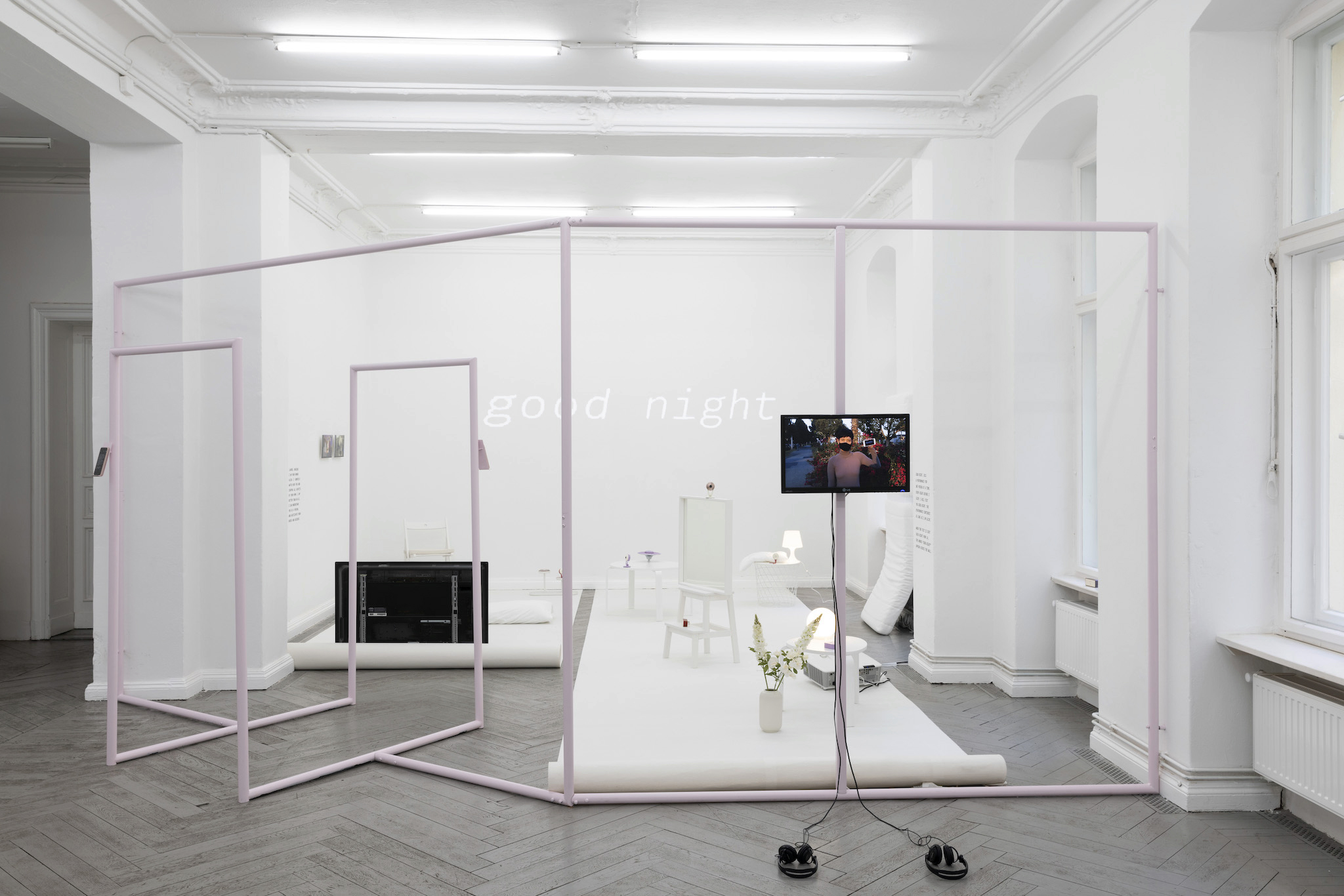 Pictured is an installation, in a brightly-lit white walled-room with a light pink minimal steel frame traces the boundary of the work, like a blueprint of a small, intimate living space with furniture such as a white wooden coffee table, mirror with a webcam, TV attached to the frame, print outs of excerpts of conversations had during the height of the COVID-19 Pandemic.