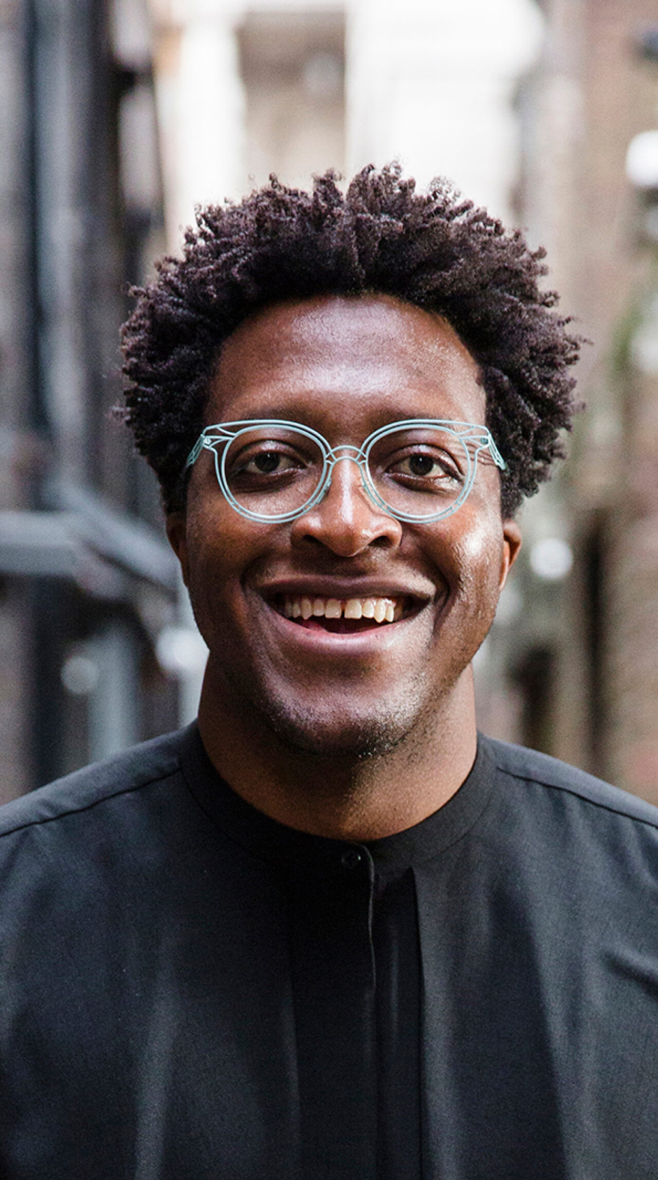 A young African man with an open mouth smile showing teeth, he's wearing a black collar-less shirt and blue glasses which contrast against his dark brown skin. He is standing in an outdoor urban space.