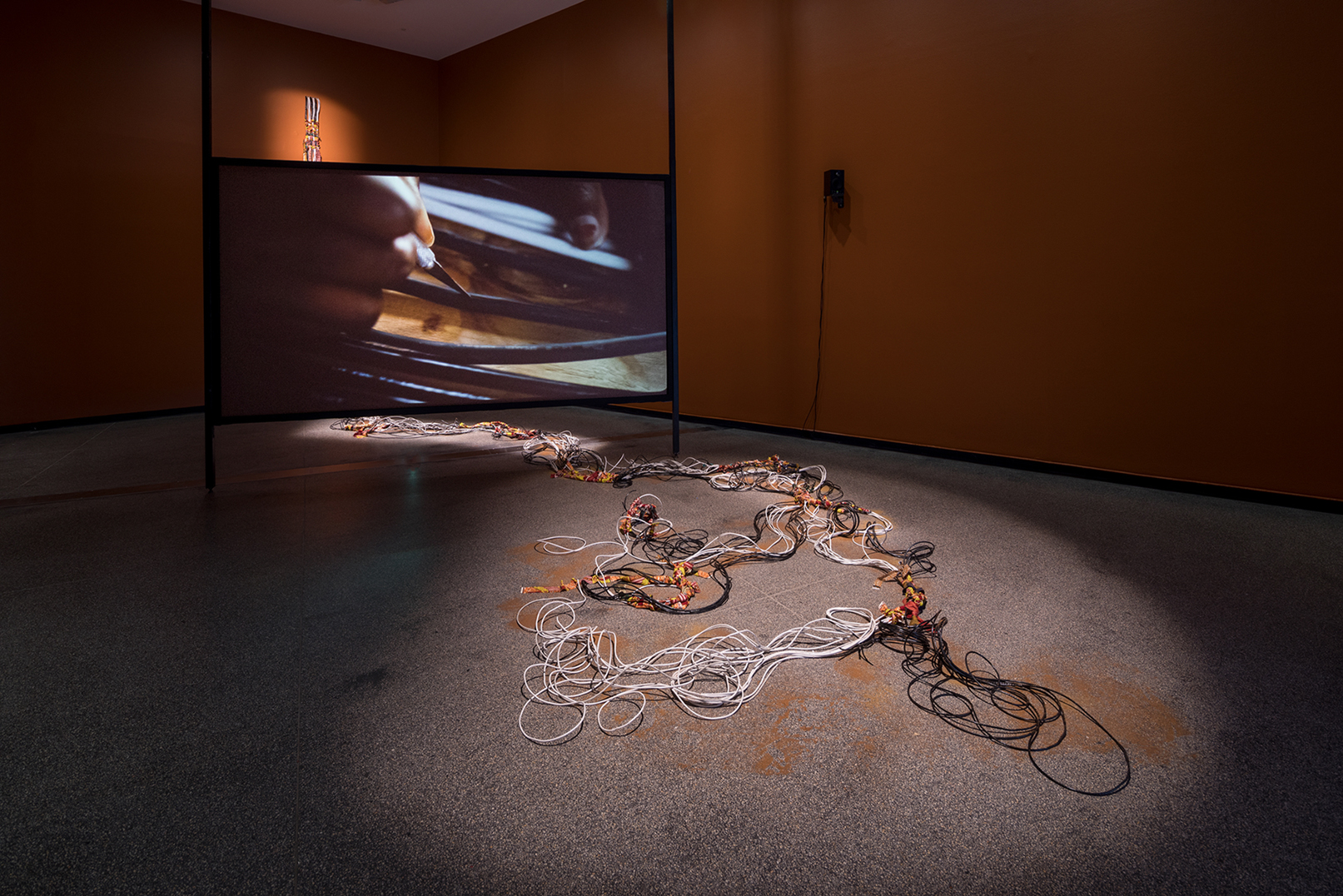 Pictured is a mixed media installation of cables, cloth, hair, dust, spices and herbs braided and intermingled together, which flows behind a screen featuring a video, onto the floor in front and loops around. Gesture of modern technical systems mixed with values and artistry and practices from other cultures.