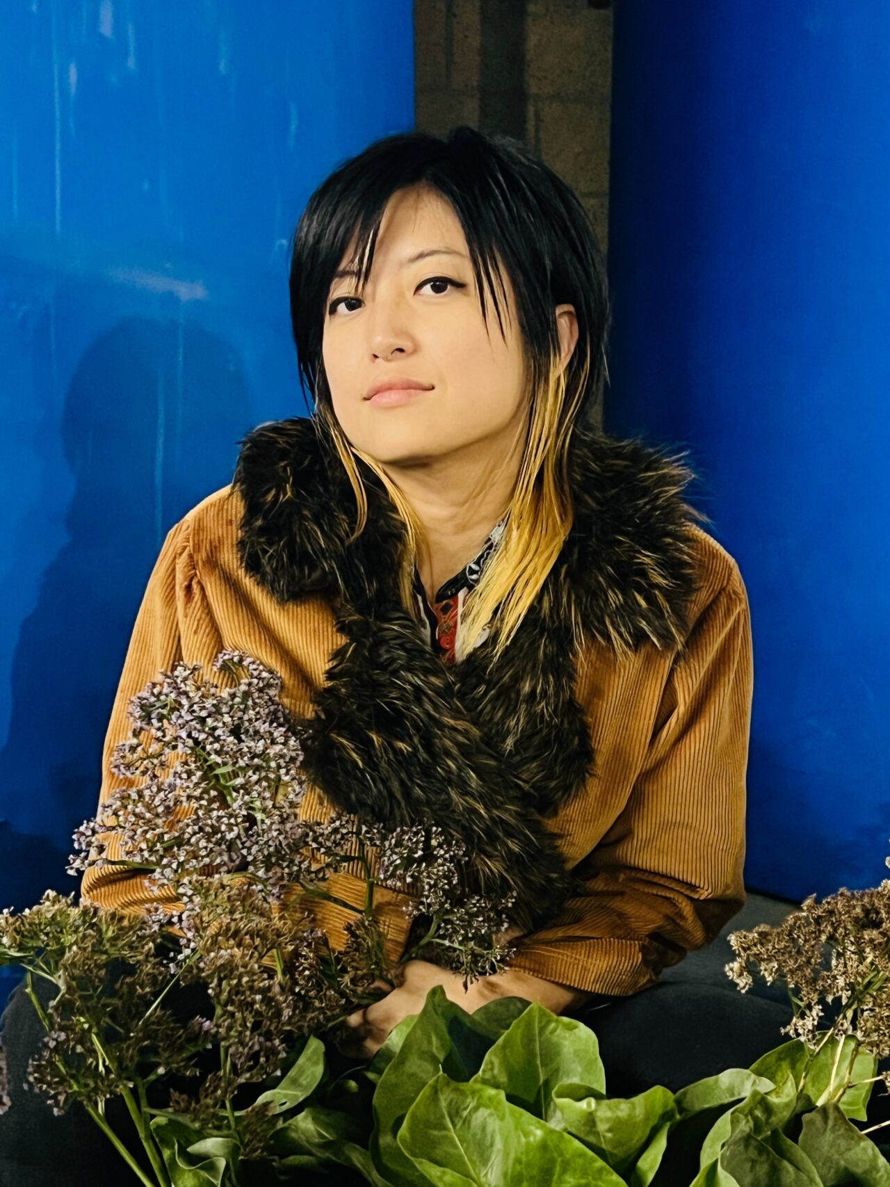 Pictured is the artist Xin Xin, an Asian-American person with a layered haircut illuminated by dark blue and warm yellow lights.