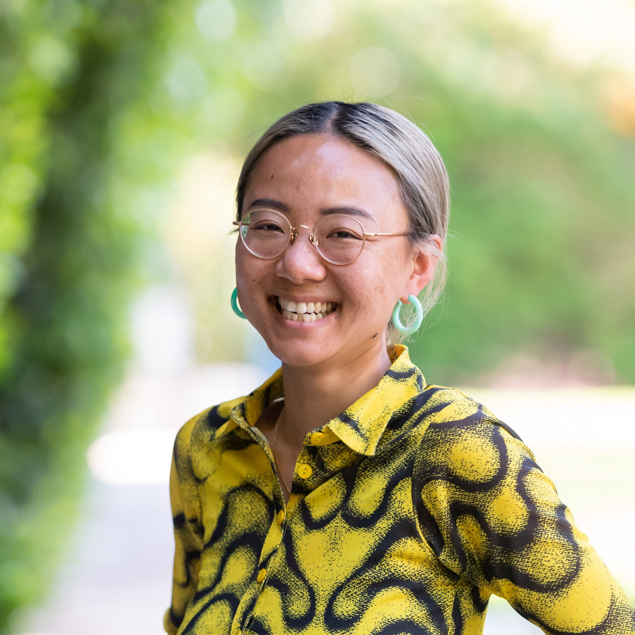 Xiaowei Wang is a Chinese person (AFAB), with dyed blond hair, and dark brown/black roots. Xiaowei is wearing a cool funky, button-up blouse with yellow and black round patterns, and jade earrings. They are standing outside, smiling brightly in front of shrubs and trees in what seems like a sunny day.